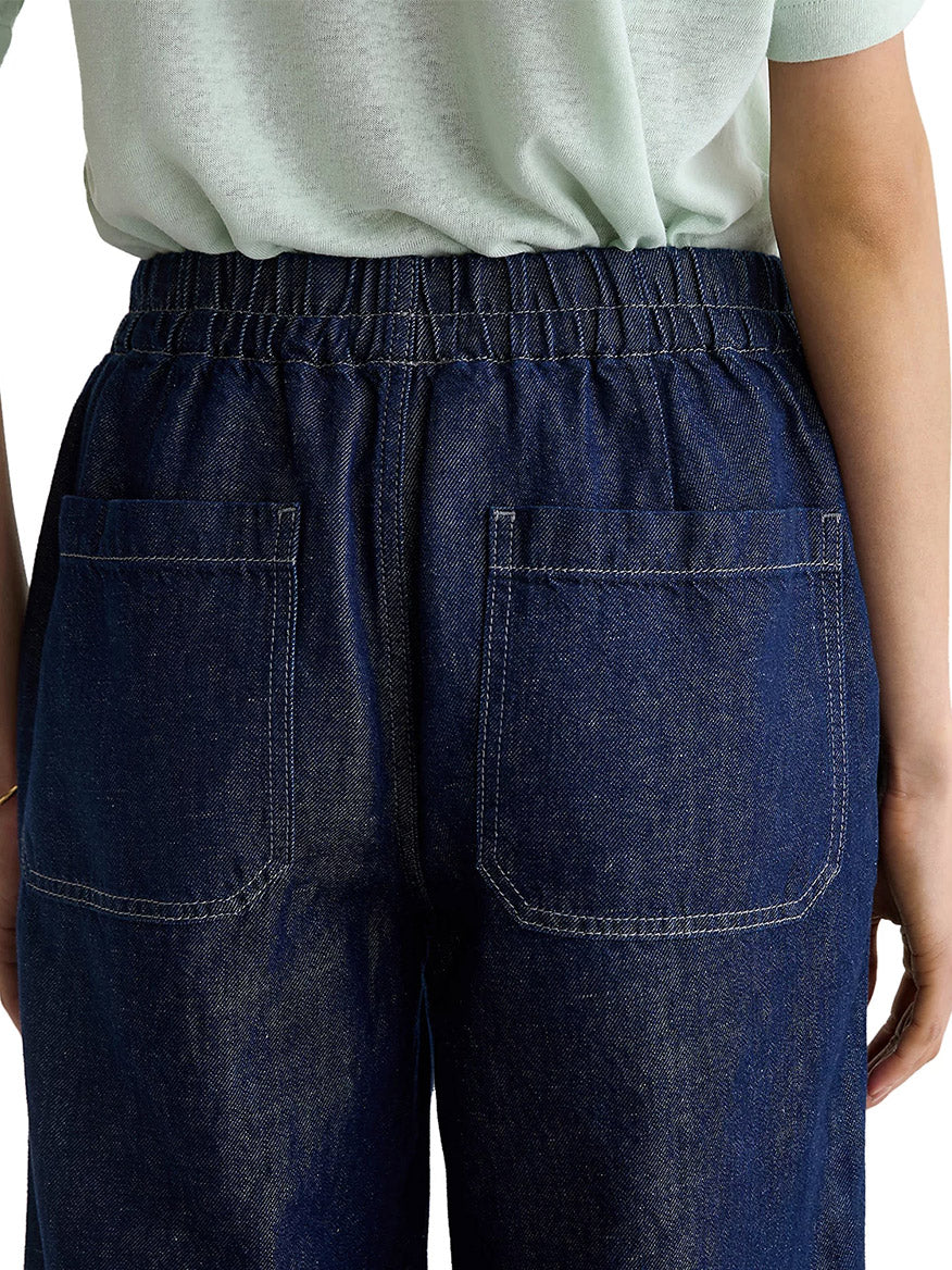 Model wearing AG Jeans Adria Cinched Jean Low-Rise Wide Leg in Napa, a dark blue linen-blend denim with an elastic waistband and two back pockets, shown from the waist down, facing away from the camera.