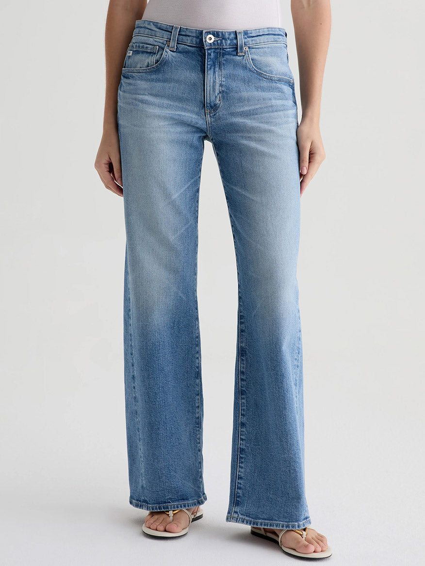 Wearing AG Jeans Adria Twisted Leg Low-Rise Wide Leg in Helios, a white top, and beige sandals, a person stands against a plain light background.