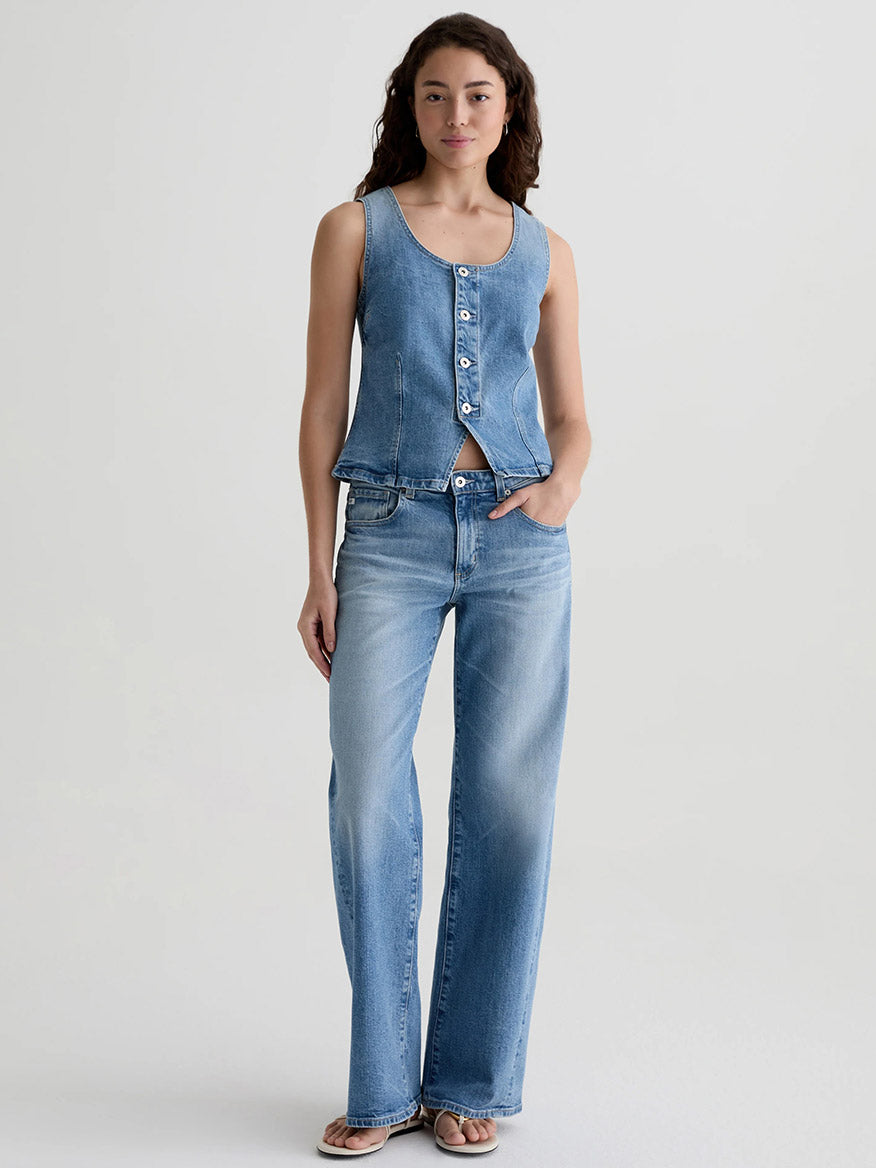 A woman stands against a plain background wearing the AG Jeans Adria Twisted Leg Low-Rise Wide Leg in Helios with a sleeveless, buttoned Italian denim top and sandals.
