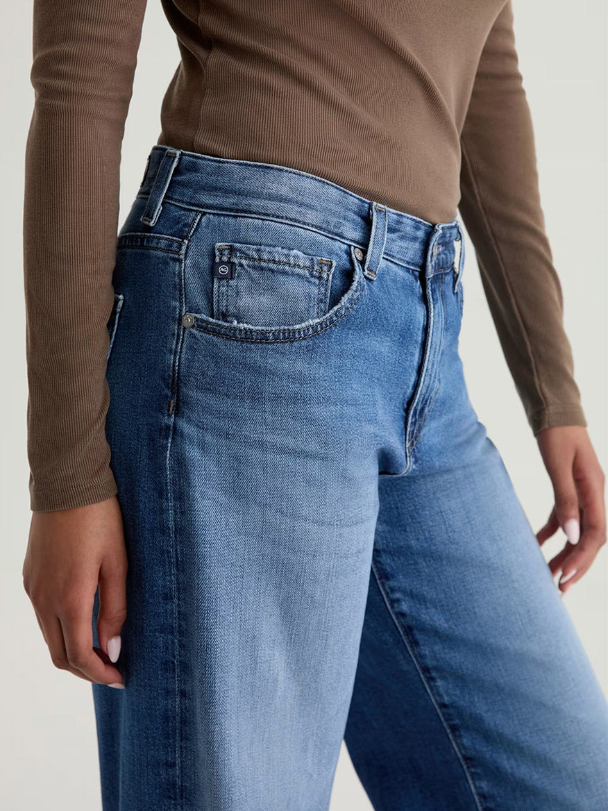 A person is wearing a brown long-sleeve top and AG Jeans Adria Low-Rise Wide Leg in Headline denim, shown from mid-torso to thigh.