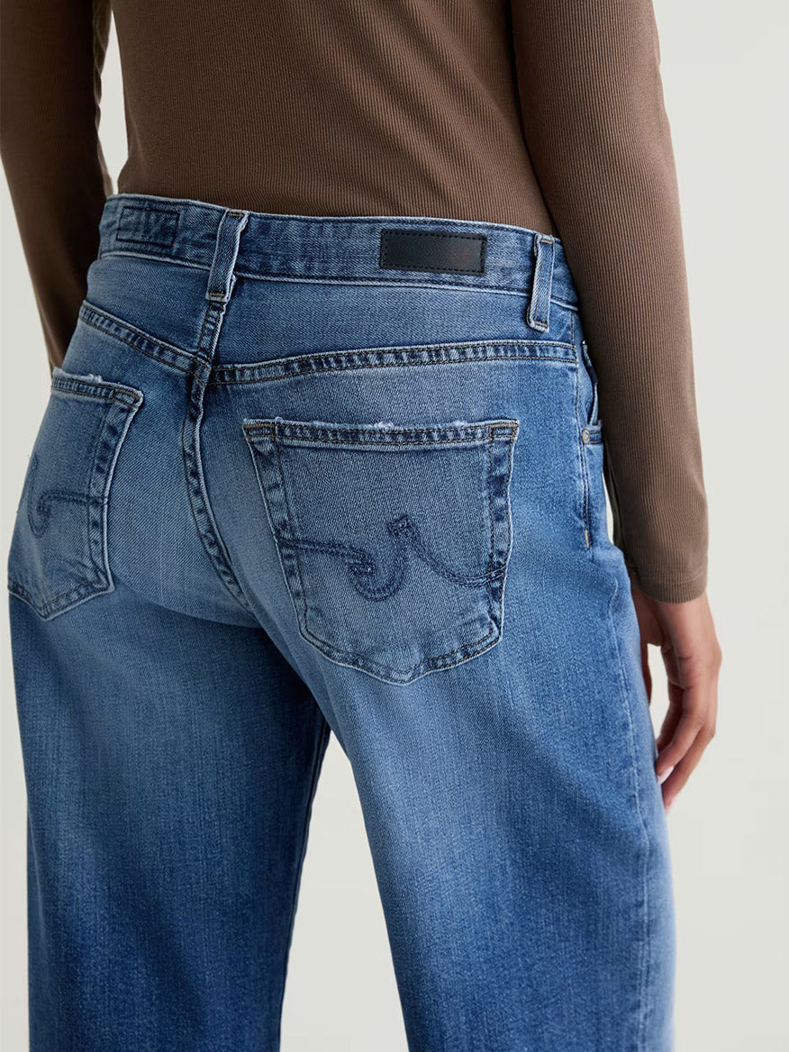 Person wearing AG Jeans Adria Low-Rise Wide Leg in Headline and a long-sleeve brown top, shown from the back. These low-rise jeans feature two back pockets with stitched designs, crafted from premium Japanese denim.