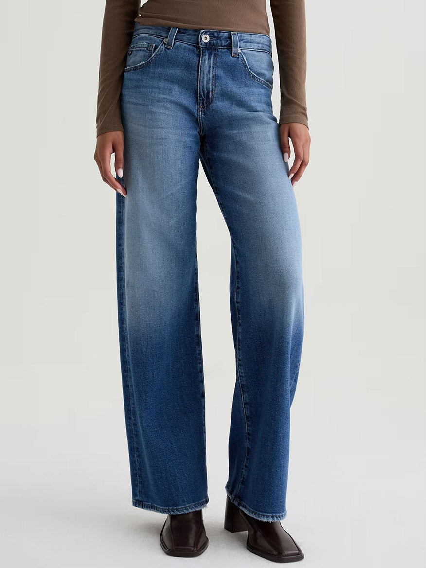 Person wearing AG Jeans Adria Low-Rise Wide Leg in Headline, premium blue Japanese denim jeans, and a brown long-sleeve top, standing against a plain light background.
