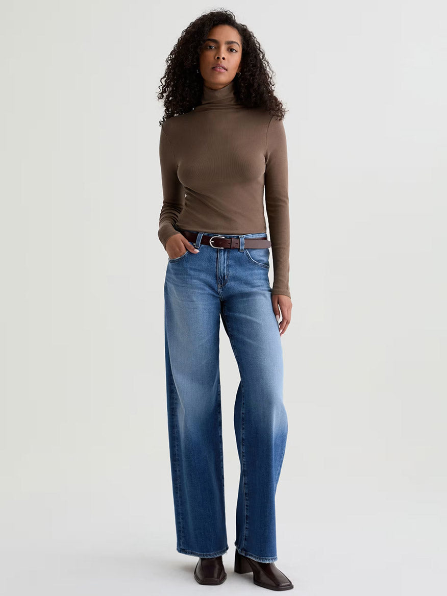 A person stands against a plain background wearing a brown turtleneck, AG Jeans Adria Low-Rise Wide Leg in Headline, a brown belt, and brown shoes.