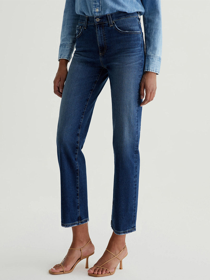 A person wearing a light blue denim shirt, AG Jeans Brinley Crop in 7 Years La Jolla, and beige strappy heeled sandals stands against a plain background.