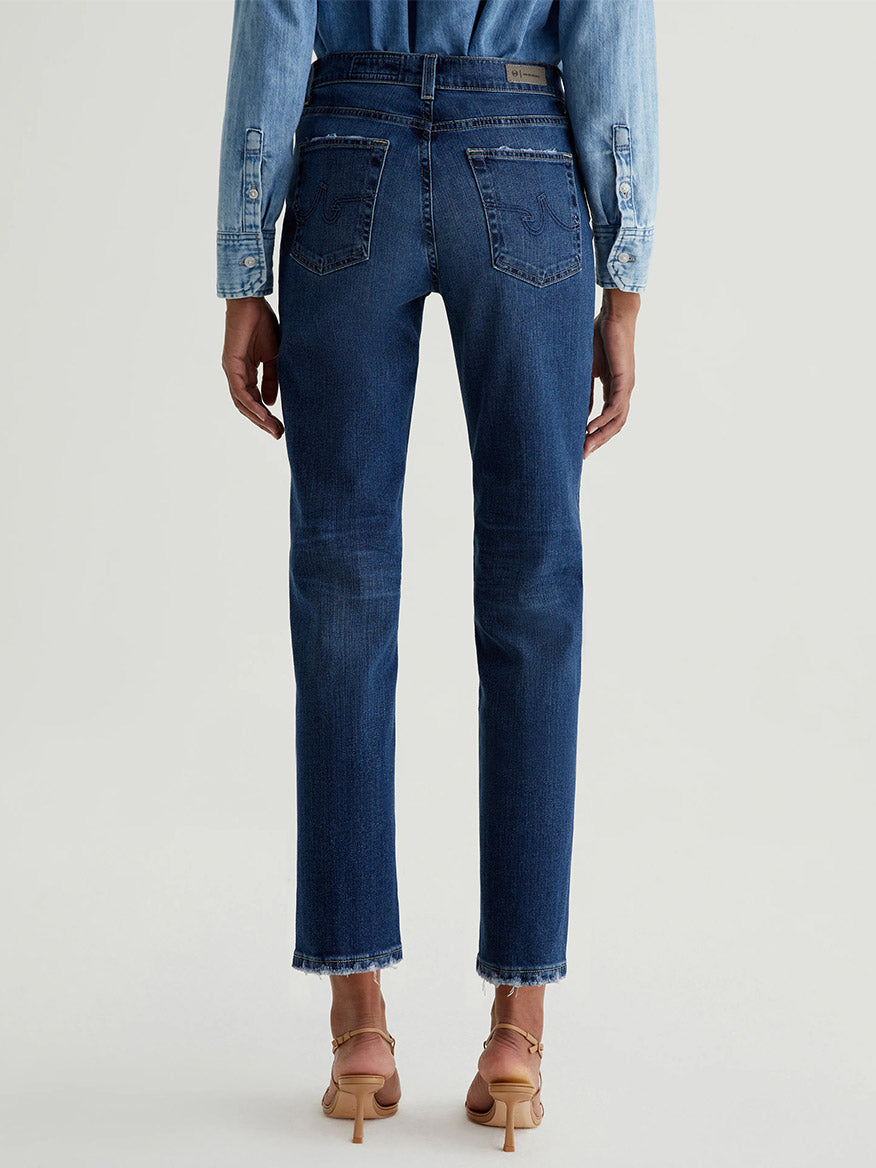 Model wears the AG Jeans Brinley Crop in 7 Years La Jolla, paired with a denim shirt and beige heeled sandals, shown from the back on a light grey background for a vintage-inspired comfort stretch denim look.