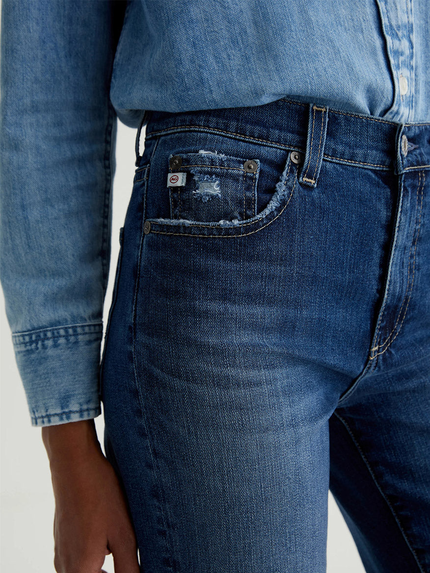 Wearing the AG Jeans Brinley Crop in 7 Years La Jolla, a person pairs a light blue denim shirt with these dark indigo, high-waisted jeans featuring vintage comfort stretch and a frayed coin pocket for modern style.