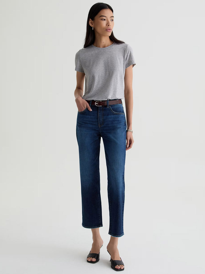 A woman stands against a plain background wearing AG Jeans Brinley Crop in Outspoken, a grey t-shirt, a brown belt, black sandals, and a bracelet on her left wrist.