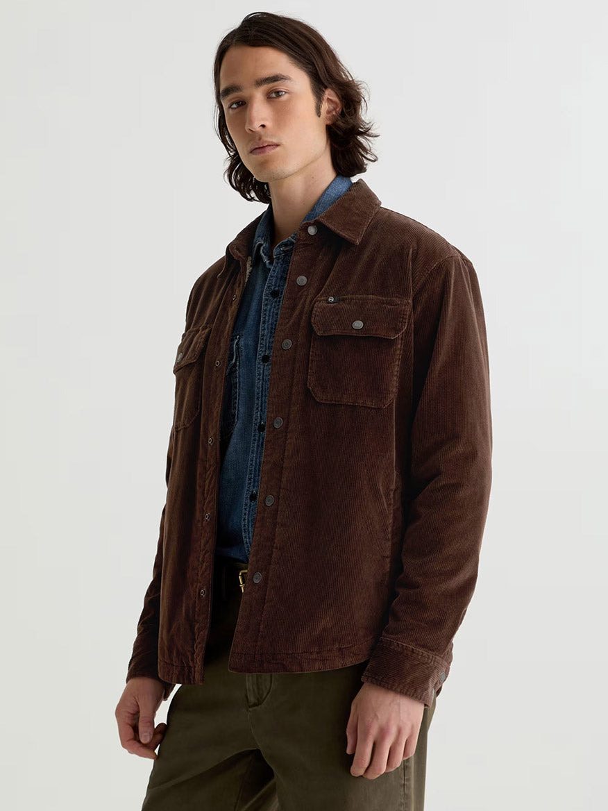 A man wears the AG Jeans Elias Corduroy Shirt Jacket in Raw Umber, styled oversized over a blue denim shirt and olive pants, standing against a plain light background.
