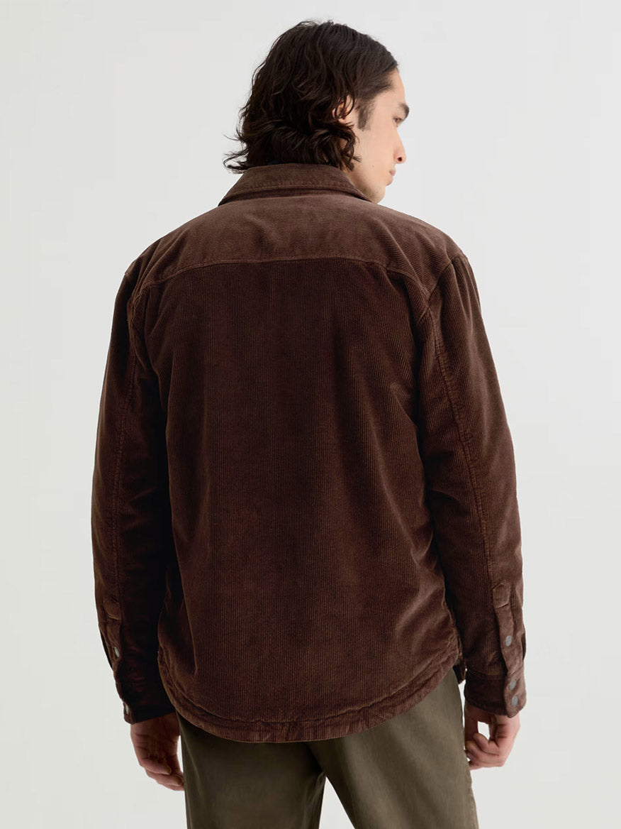 A person with medium-length dark hair wears the AG Jeans Elias Corduroy Shirt Jacket in Raw Umber and olive pants, standing with their back to the camera against a plain light background.