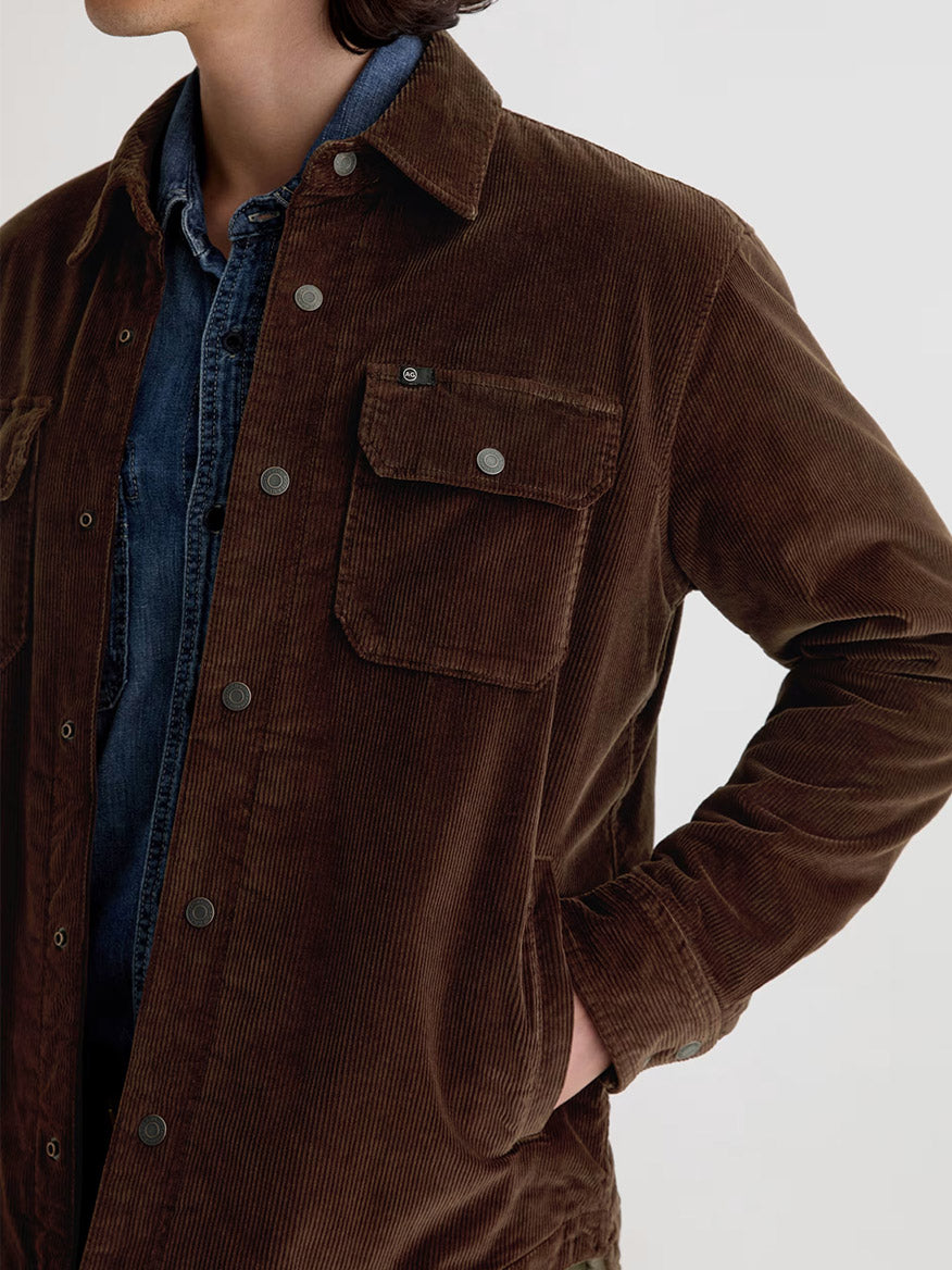 A person wears the AG Jeans Elias Corduroy Shirt Jacket in Raw Umber over a blue denim shirt, with one hand in their pocket, shown from shoulders to waist.