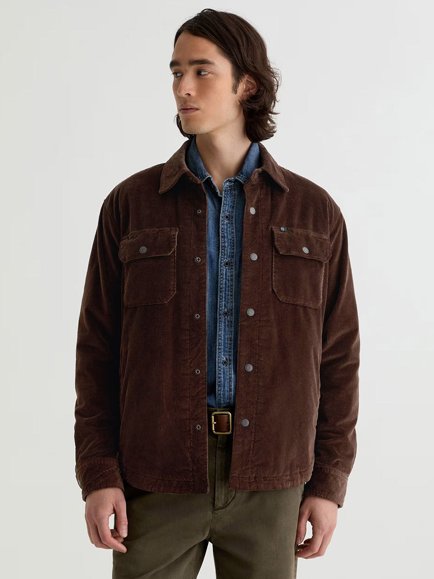 A man wears the AG Jeans Elias Corduroy Shirt Jacket in Raw Umber over a denim shirt with olive green pants, standing against a plain light background.