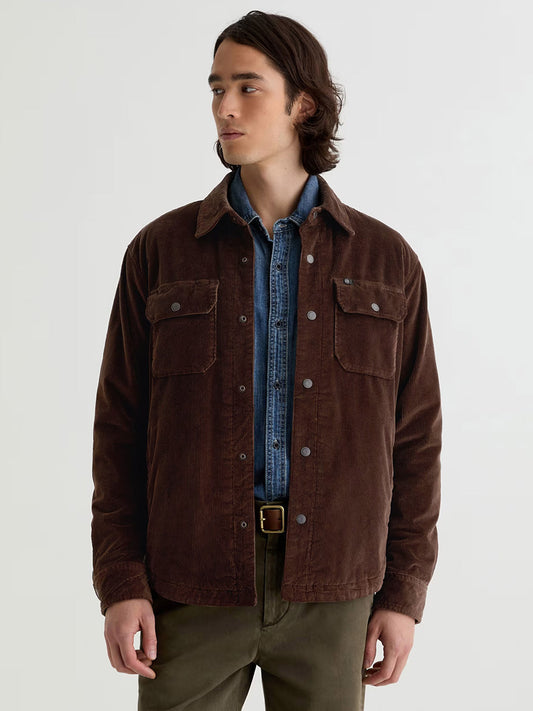 A man wears the AG Jeans Elias Corduroy Shirt Jacket in Raw Umber over a denim shirt with olive green pants, standing against a plain light background.