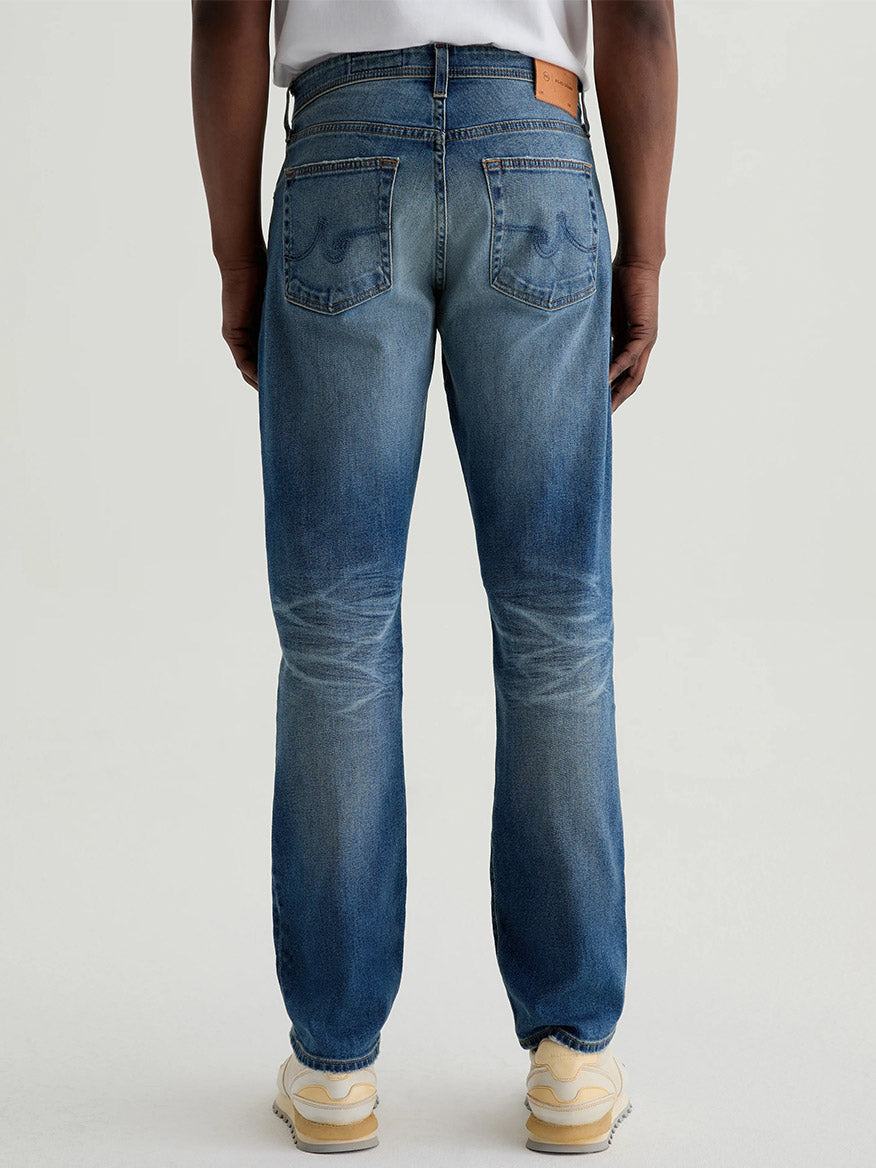 A person stands facing away against a plain background, wearing AG Jeans Everett in 13 Years San Martin—medium blue slim straight jeans with back pockets and a brown patch—paired with a tucked-in white shirt and light sneakers.