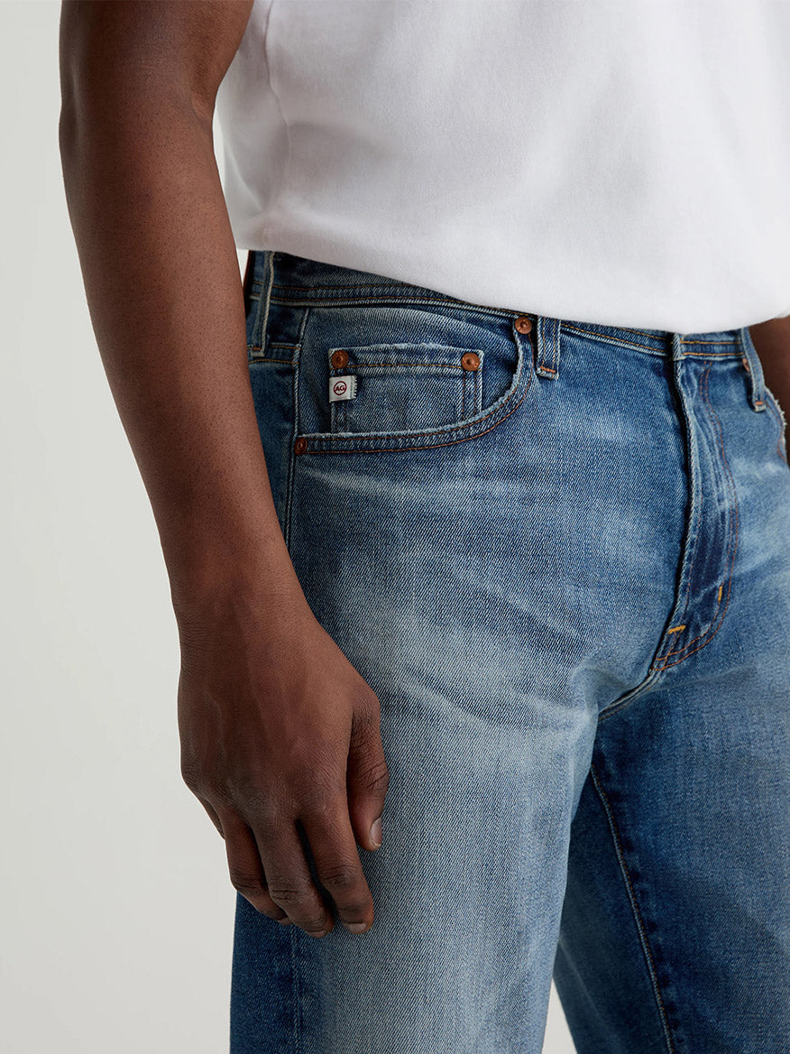 Wearing AG Jeans Everett in 13 Years San Martin, a person stands with their left arm at their side. The white shirt and medium blue slim straight jeans are visible from neck to mid-thigh in the cropped image.