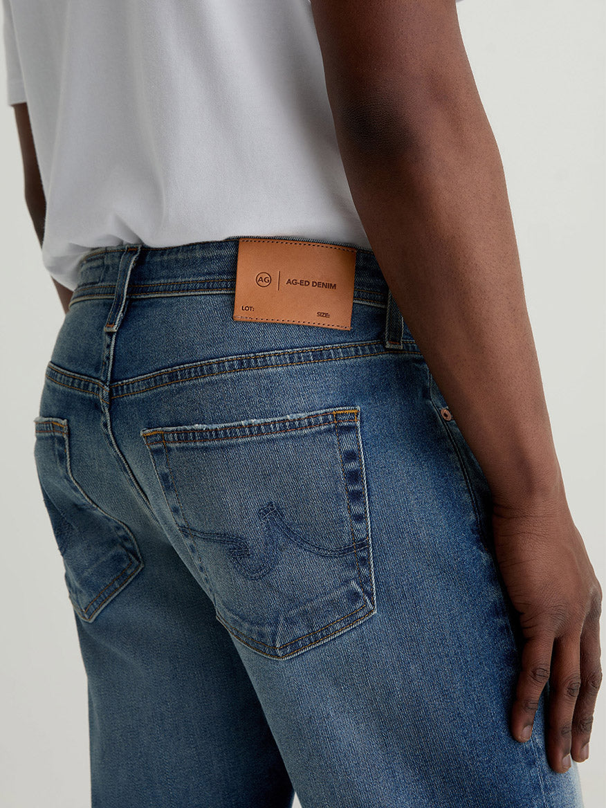 A person wearing AG Jeans Everett in 13 Years San Martin, a slim straight medium blue jean, with a white shirt and a brown leather label on the waistband reading "Aged Denim," shown from the waist down.