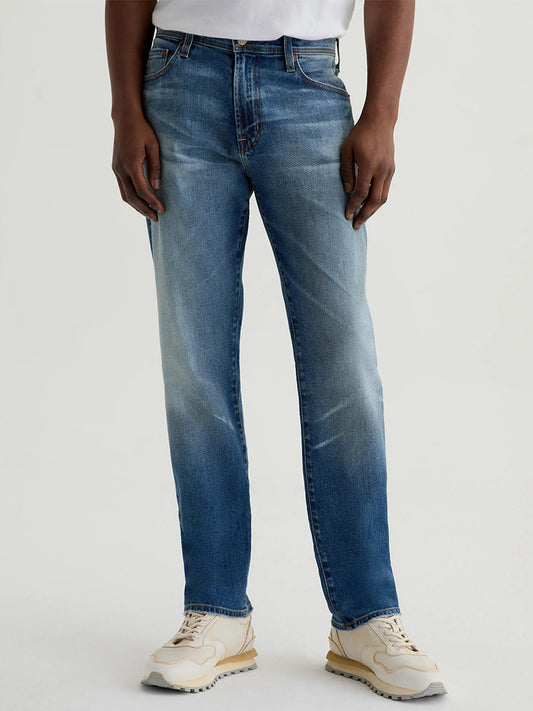 A person wearing a white shirt, white sneakers, and AG Jeans Everett in 13 Years San Martin stands against a plain light background.