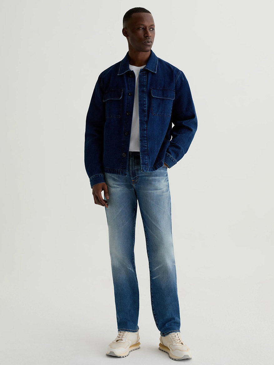 A man stands against a plain background wearing the AG Jeans Everett in 13 Years San Martin—medium blue, slim straight jeans crafted from Japanese comfort stretch denim—paired with a dark denim jacket, white t-shirt, and beige sneakers.