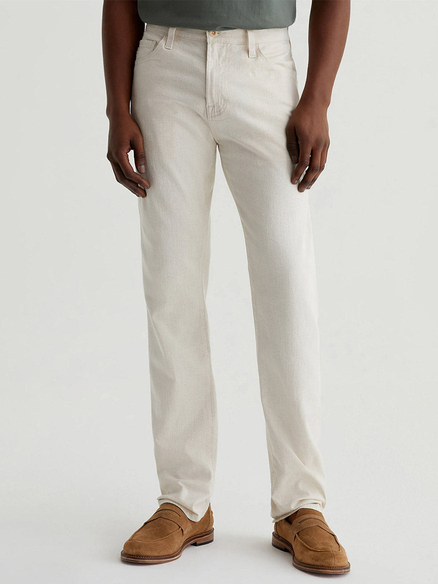 Person wearing AG Jeans Everett in Natural—mid-rise, light-colored slim straight jeans—with a green shirt and brown loafers, standing against a plain light background.
