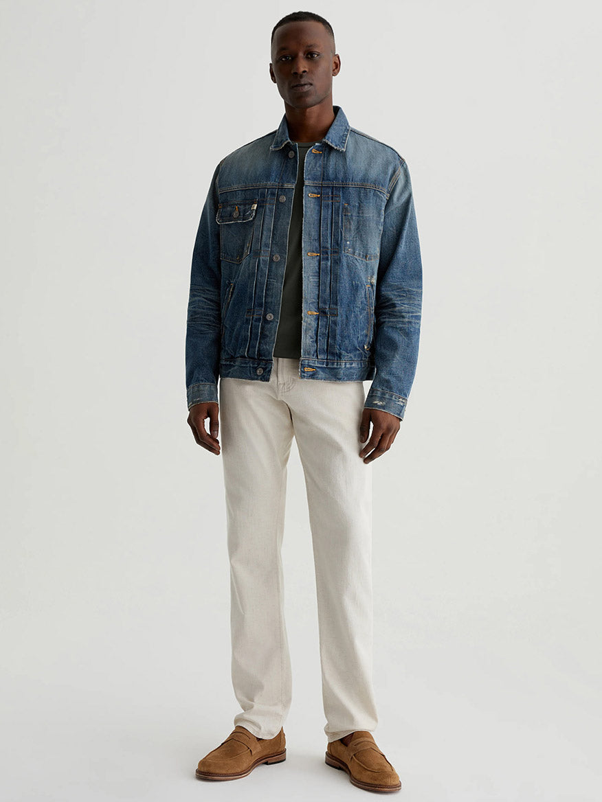 A man stands against a plain background wearing a blue denim jacket, dark shirt, AG Jeans Everett in Natural linen pants, and brown shoes.