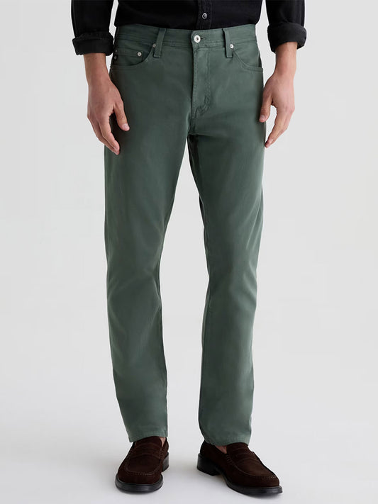 Someone wearing AG Jeans Everett Sueded in Cypress Green, a black long-sleeve shirt, and brown loafers stands against a plain light background.