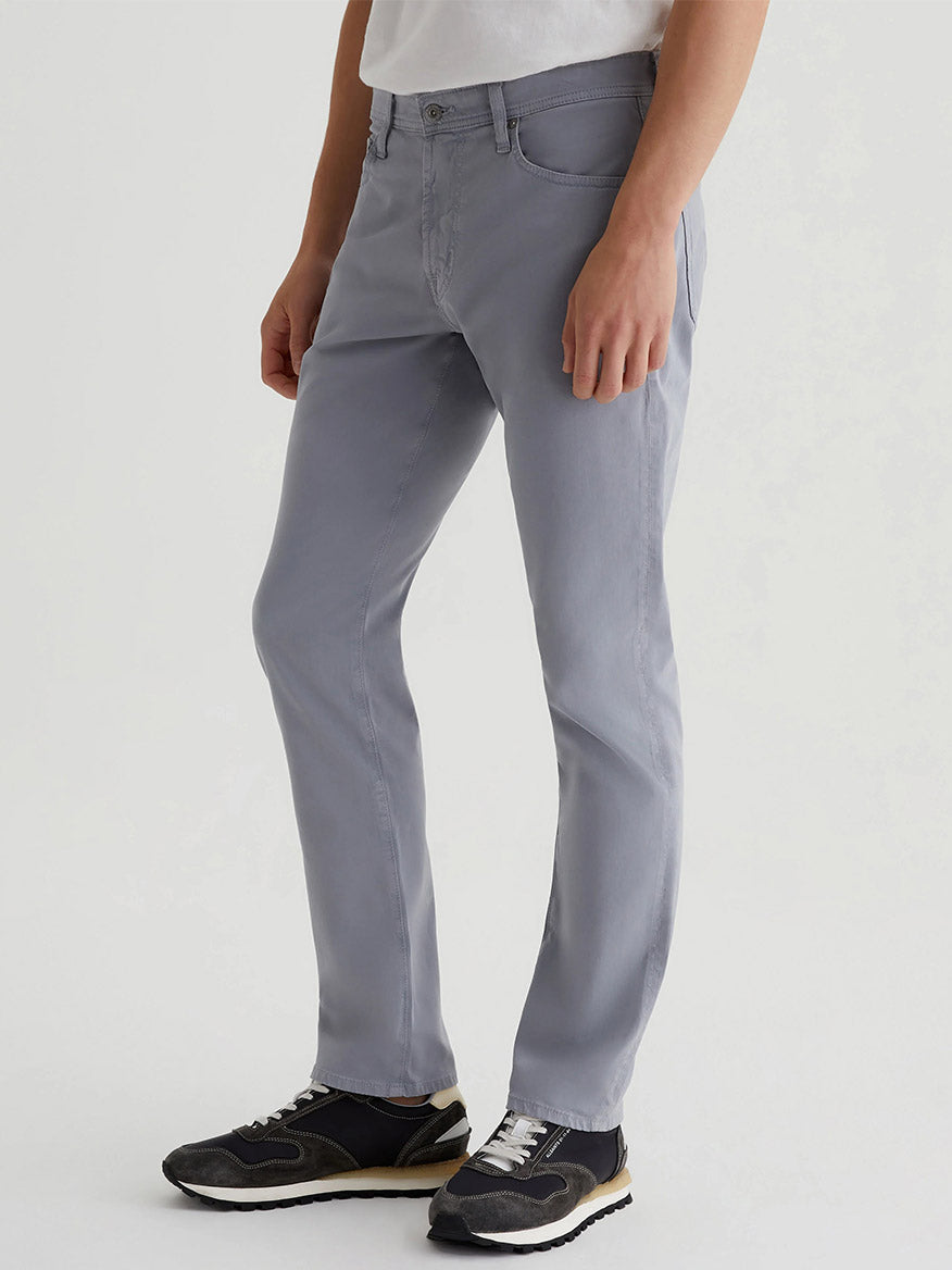 A person in gray skinny jeans, a white shirt, and black sneakers stands against a plain light background, embodying the effortless style of AG Jeans Everett Sueded in Ocean Breeze.