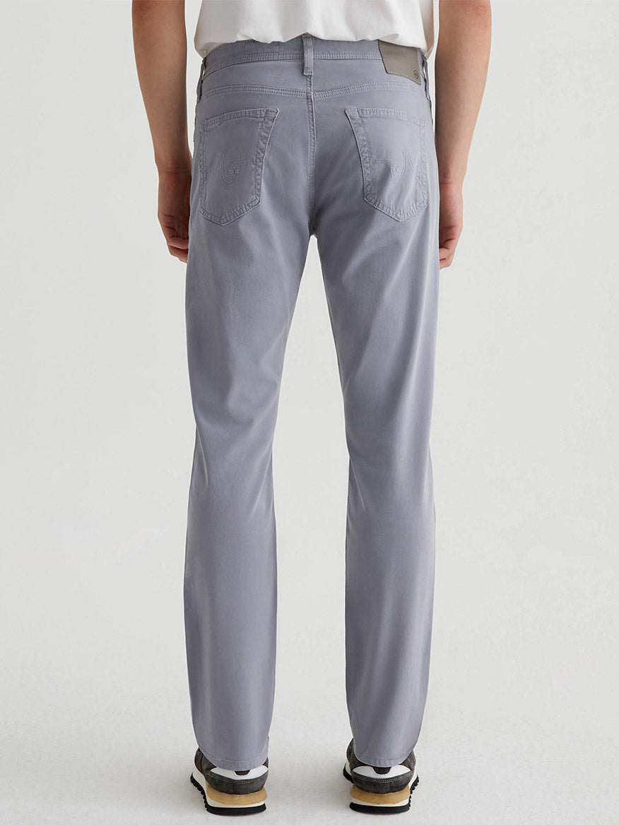A person wearing AG Jeans Everett Sueded in Ocean Breeze, along with a white shirt and black sneakers, stands against a plain background.