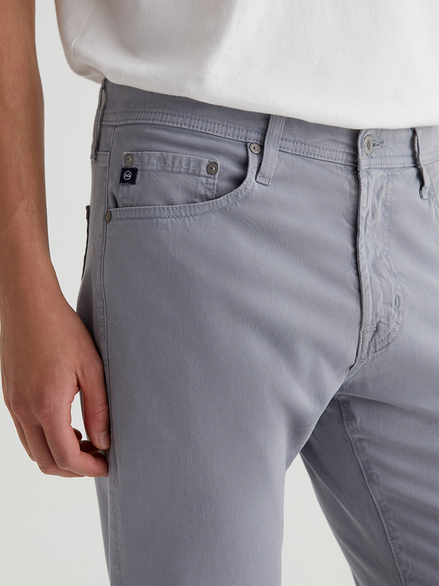 Person in light gray AG Jeans Everett Sueded in Ocean Breeze with a white top. Visible side pocket, buttons, and belt loops enhance the style. Jeans are fitted yet casual, ideal for a relaxed day out.