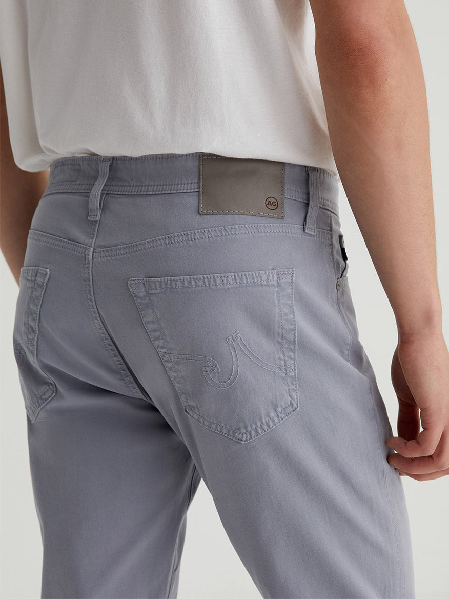Viewed from behind, a person wears light gray AG Jeans Everett Sueded in Ocean Breeze and a white shirt. Crafted from Sueded Stretch Sateen, these slim-straight jeans boast curved stitching on the back pocket for subtle flair.