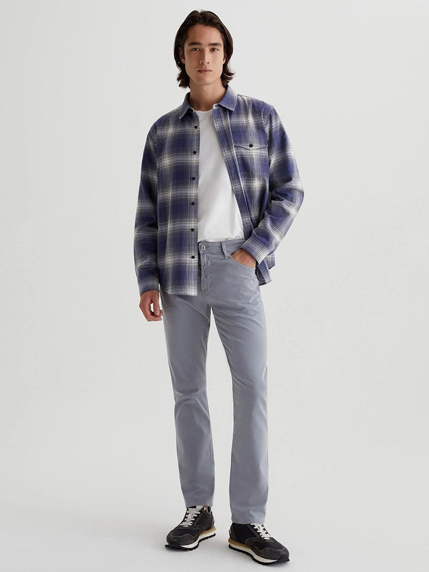 A person in a blue plaid shirt over a white T-shirt, AG Jeans Everett Sueded in Ocean Breeze gray pants, and black sneakers stands against a plain background.