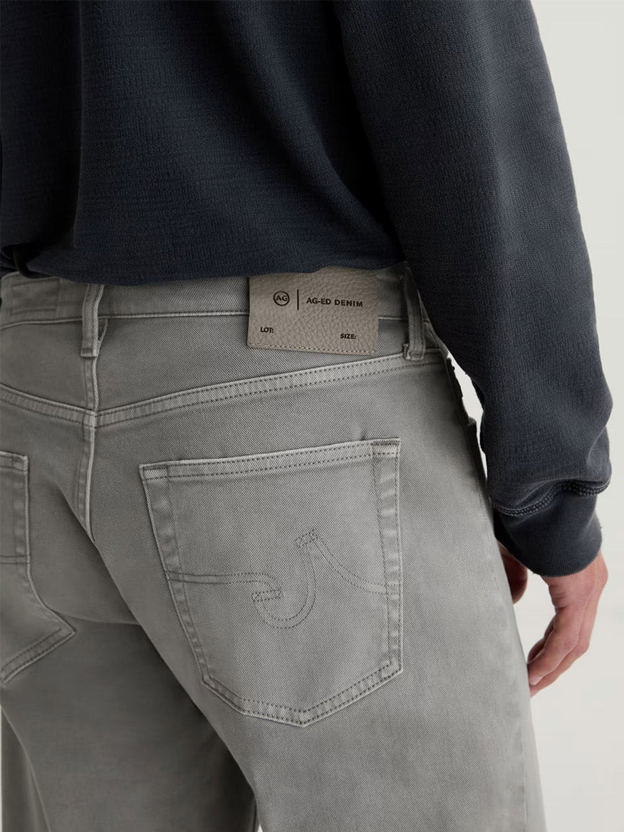 Person wearing AG Jeans Everett in 7 Years Sulfur Ironwood—a slim straight fit in a cold grey wash—with a dark sweatshirt and the beige denim brand label visible on the back waistband.