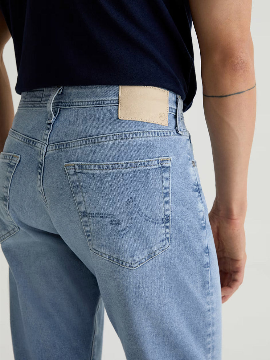Wearing AG Jeans Everett in Saltillo, a slim straight light blue jean, paired with a navy shirt; the back pocket features a stitched design and a visible beige label on the waistband.