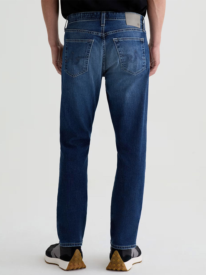A person wearing AG Jeans Everett in Upton—men’s slim straight premium denim—paired with a black shirt and black sneakers with white soles, shown from the back against a plain background.