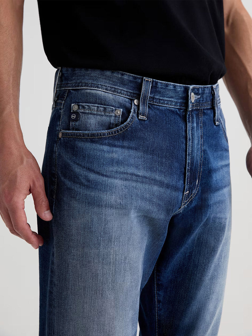 A person wearing AG Jeans Everett in Upton, a men’s slim straight jean, paired with a black t-shirt, shown from waist to mid-thighs with one hand visible at their side.