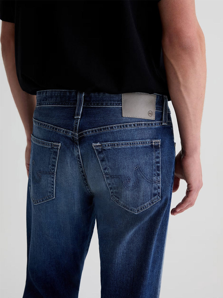 A person shown from the back wears AG Jeans Everett in Upton, a men’s slim straight premium denim, paired with a black shirt and featuring a light grey patch on the waistband.