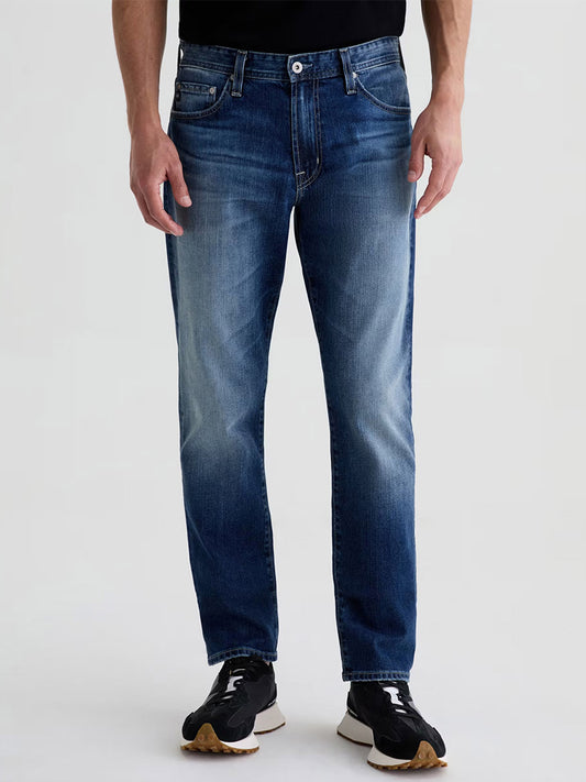 A person wearing AG Jeans Everett in Upton—men’s slim straight mid-wash blue comfort stretch denim—paired with black sneakers with white soles and a black shirt, stands against a plain light background.