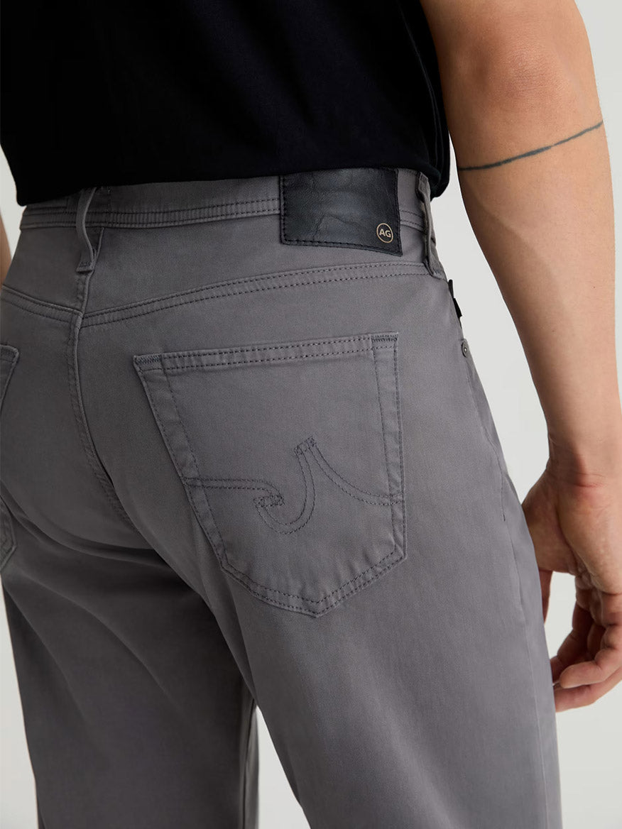 Model wearing AG Jeans Everett Sueded in Folkestone Grey, slim straight fit men's jeans with gray stretch sateen, a black shirt, and featuring stitched back pocket and small logo patch on the waistband.
