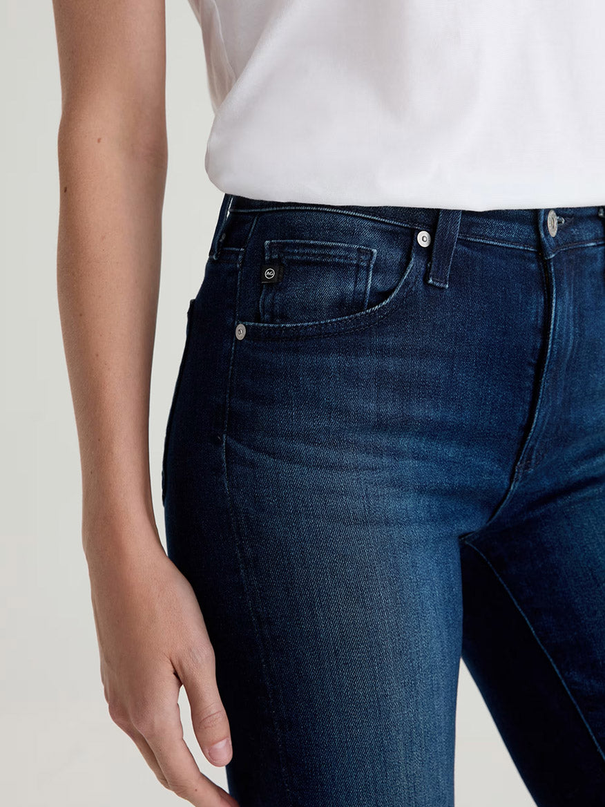 A person is shown from waist to mid-thigh, wearing a white shirt and AG Jeans Farrah Mid-Rise Boot in Epilogue—dark blue women’s bootcut jeans made from stretch denim—with one arm relaxed by their side.