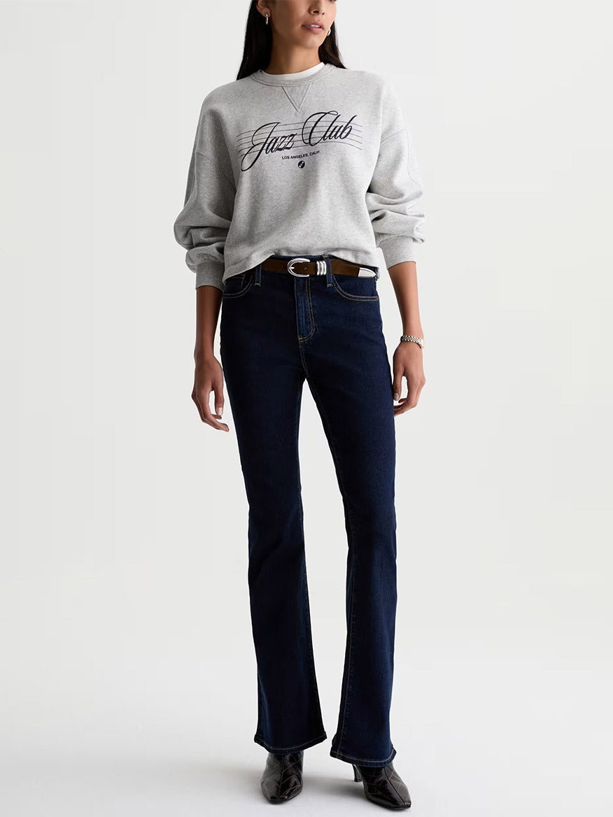 A person wearing a gray “Jazz Club” sweatshirt, a pair of AG Jeans Farrah Mid-Rise Boot in Modern Indigo, a brown belt, and dark shoes stands against a plain white background.
