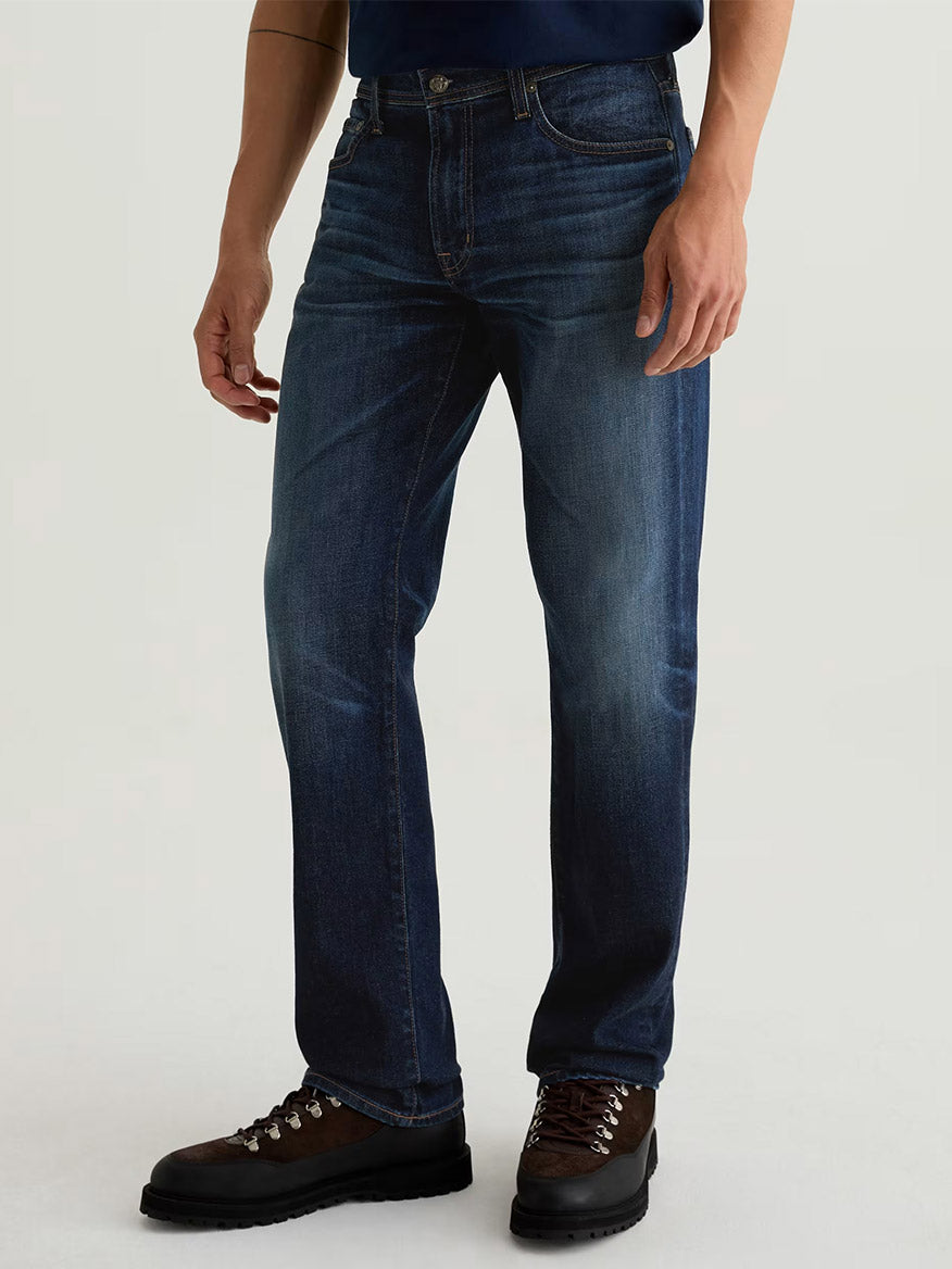 A person wearing AG Jeans Graduate in 4 Years Martel, a navy shirt, and brown hiking boots stands against a plain light background.