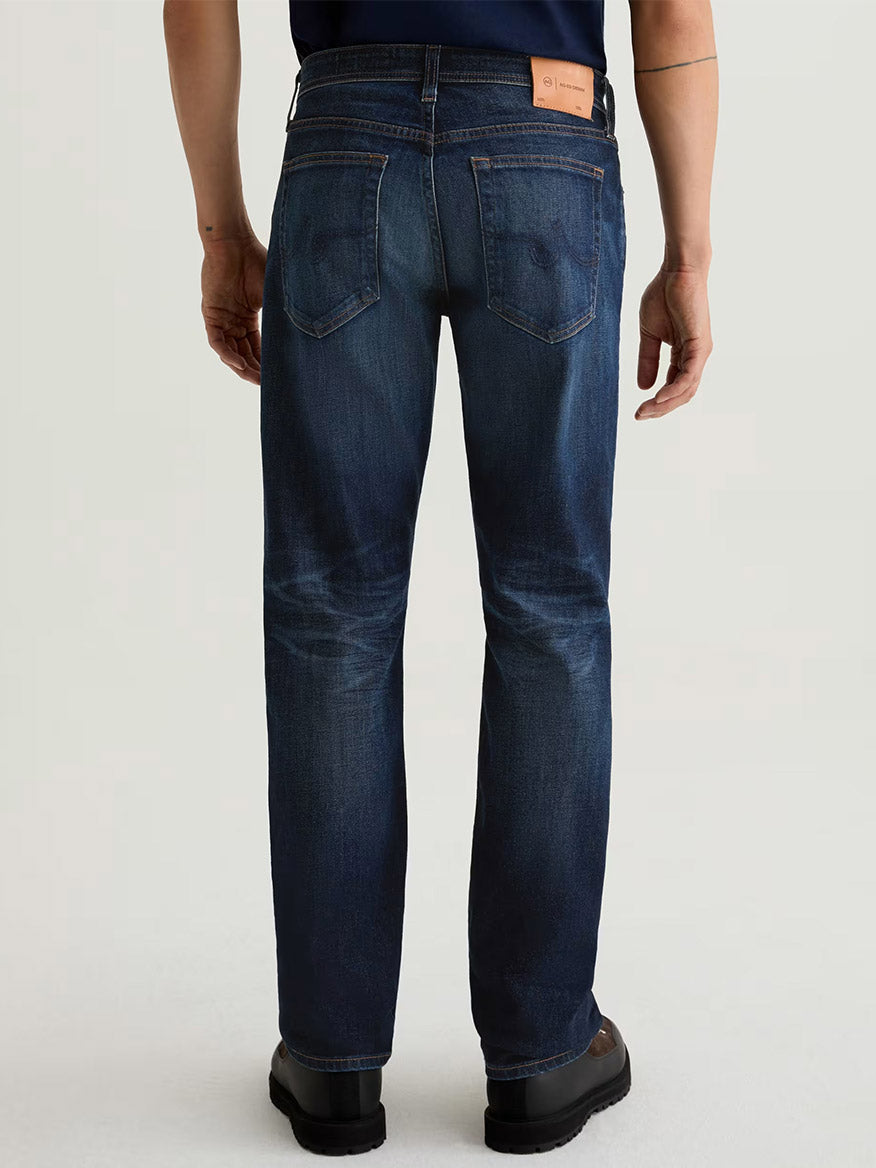 Someone wearing AG Jeans Graduate in 4 Years Martel—dark blue, relaxed fit, comfort stretch denim—is seen from the back against a plain light background.