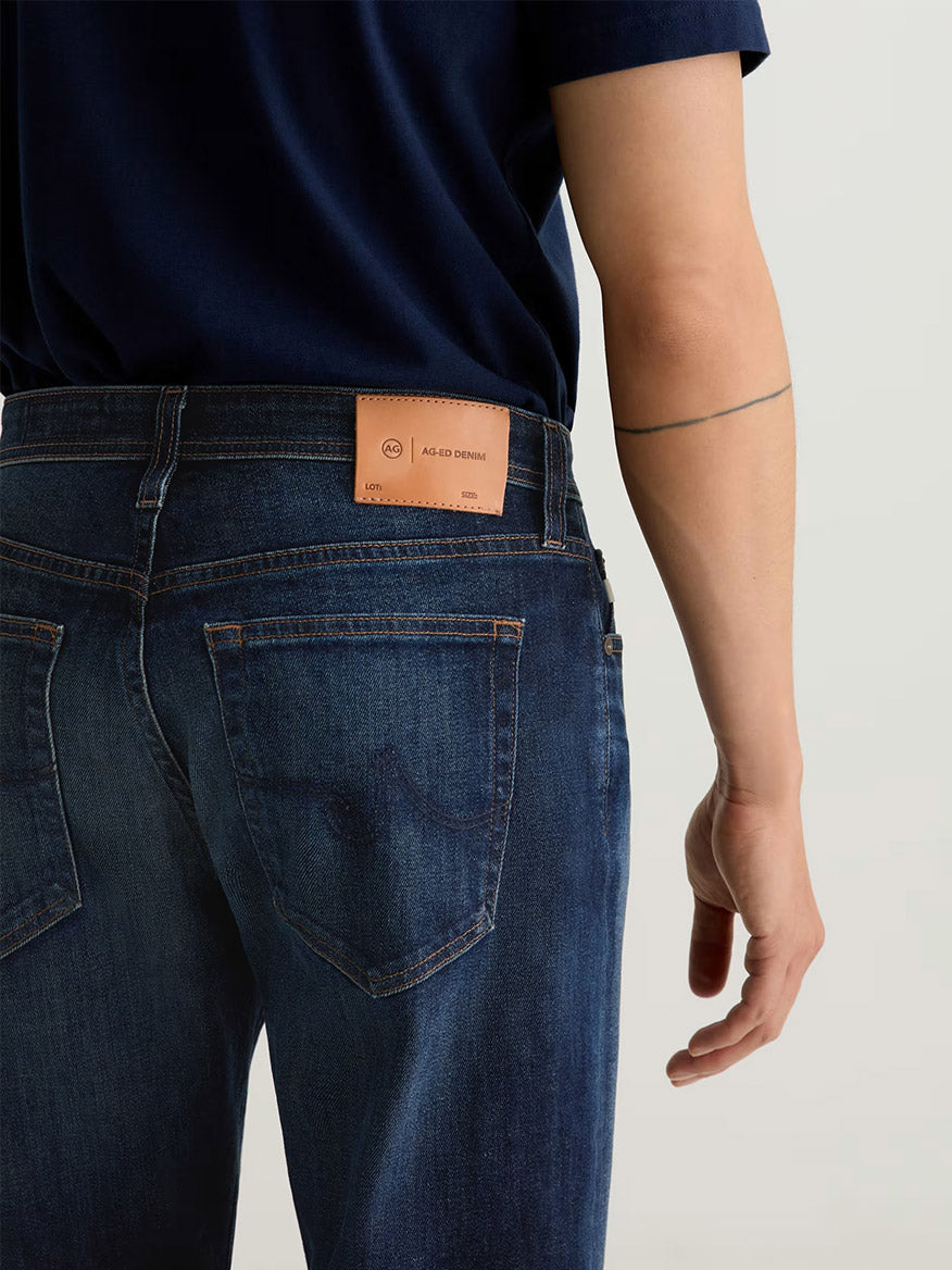 Model wears AG Jeans Graduate in 4 Years Martel, a men’s dark blue comfort stretch denim, paired with a navy shirt. Shown from the back, jeans feature a tan leather patch at the waistband and a visible arm tattoo.