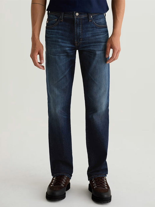 A person wears AG Jeans Graduate in 4 Years Martel—dark blue, relaxed fit, straight-leg men’s jeans—with brown hiking boots, standing against a plain light background.
