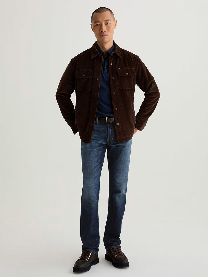 A man faces forward wearing a dark brown jacket, navy shirt, and AG Jeans Graduate in 4 Years Martel—tailored leg jeans crafted from comfort stretch denim—with brown boots against a plain light background.