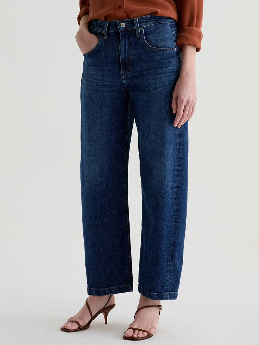 Person wearing AG Jeans Hattie High Rise Barrel in Vapor Wash Dynasty, a brown long-sleeve shirt, and brown heeled sandals, standing with one hand in their pocket against a plain background.