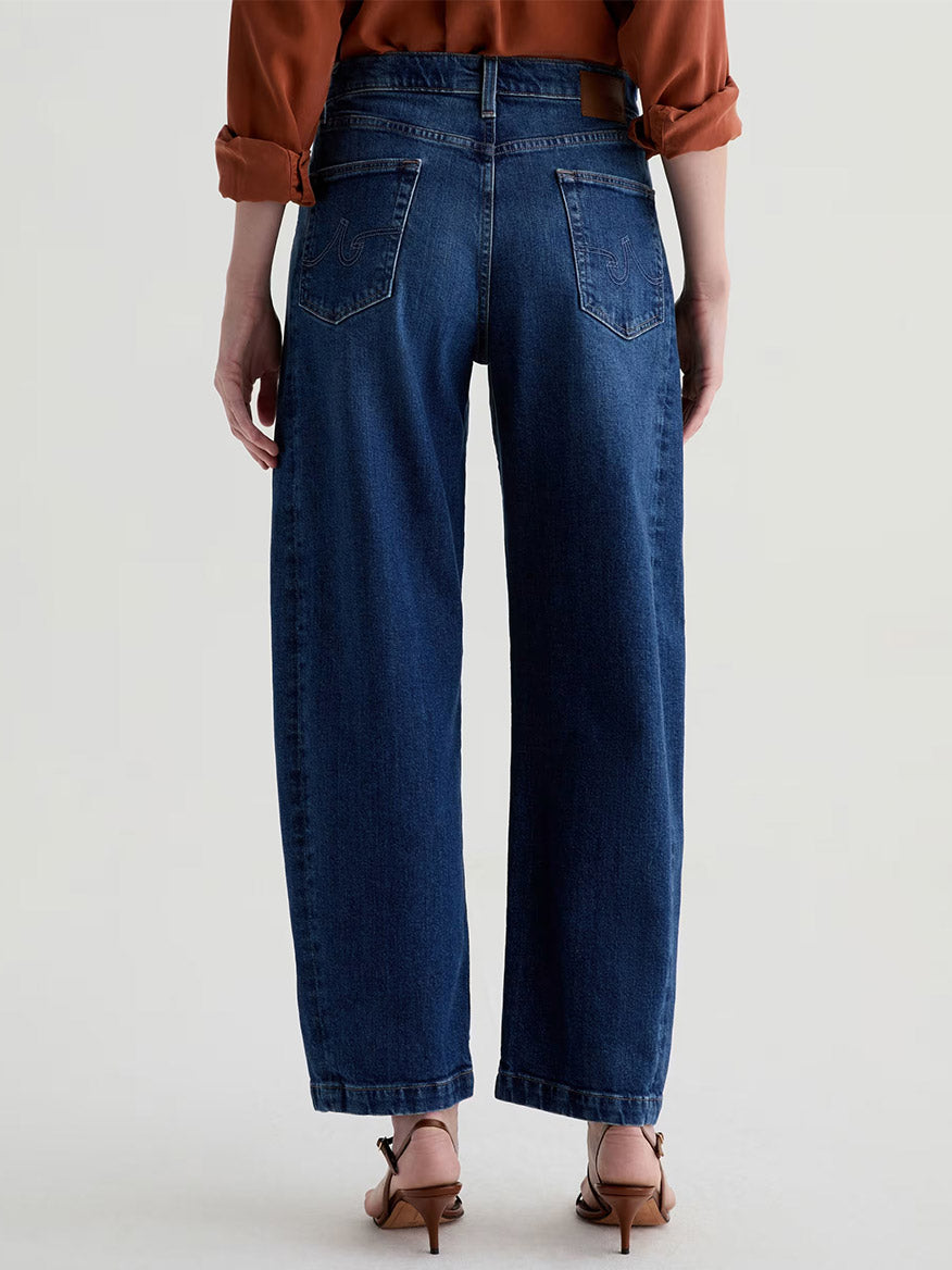 A person shown from the back wearing blue AG Jeans Hattie High Rise Barrel in Vapor Wash Dynasty, brown heeled sandals, and a rust-colored long-sleeve top.