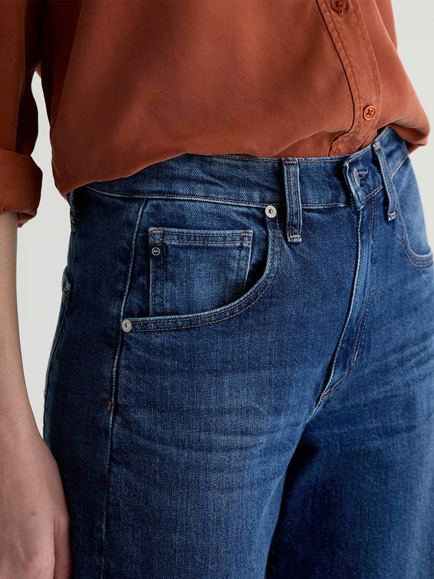 A person wears a rust-colored long-sleeve shirt tucked into AG Jeans Hattie High Rise Barrel in Vapor Wash Dynasty, shown from mid-torso to upper thigh.