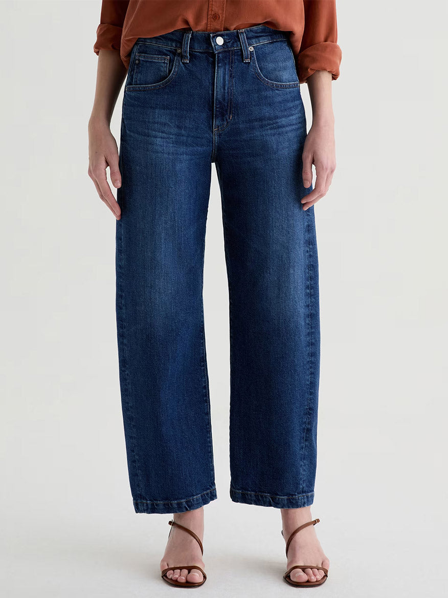 Wearing AG Jeans Hattie High Rise Barrel in Vapor Wash Dynasty, a rust long-sleeve shirt, and brown sandals, a person stands against a plain background.