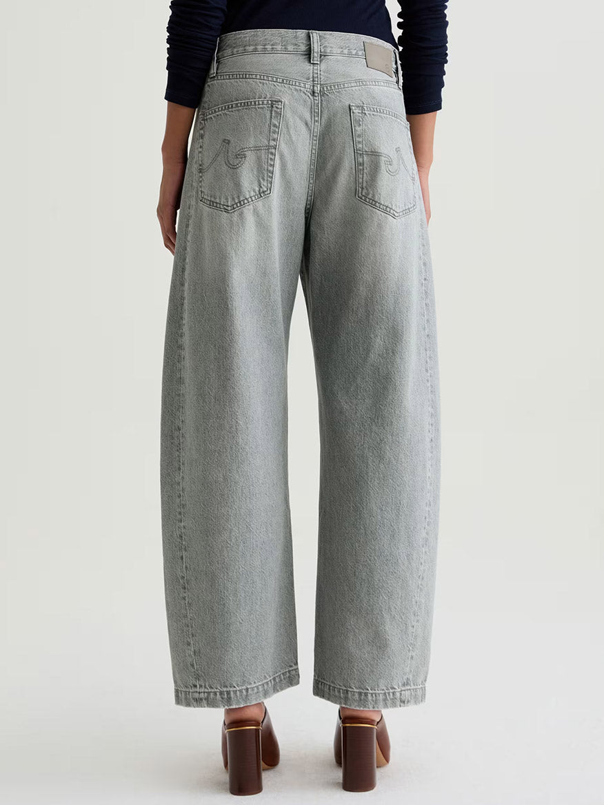 Model wearing AG Jeans Hattie High Rise Barrel in Al Fresco, a light gray non-stretch denim jean, paired with brown heeled shoes, shown from the back against a plain background.