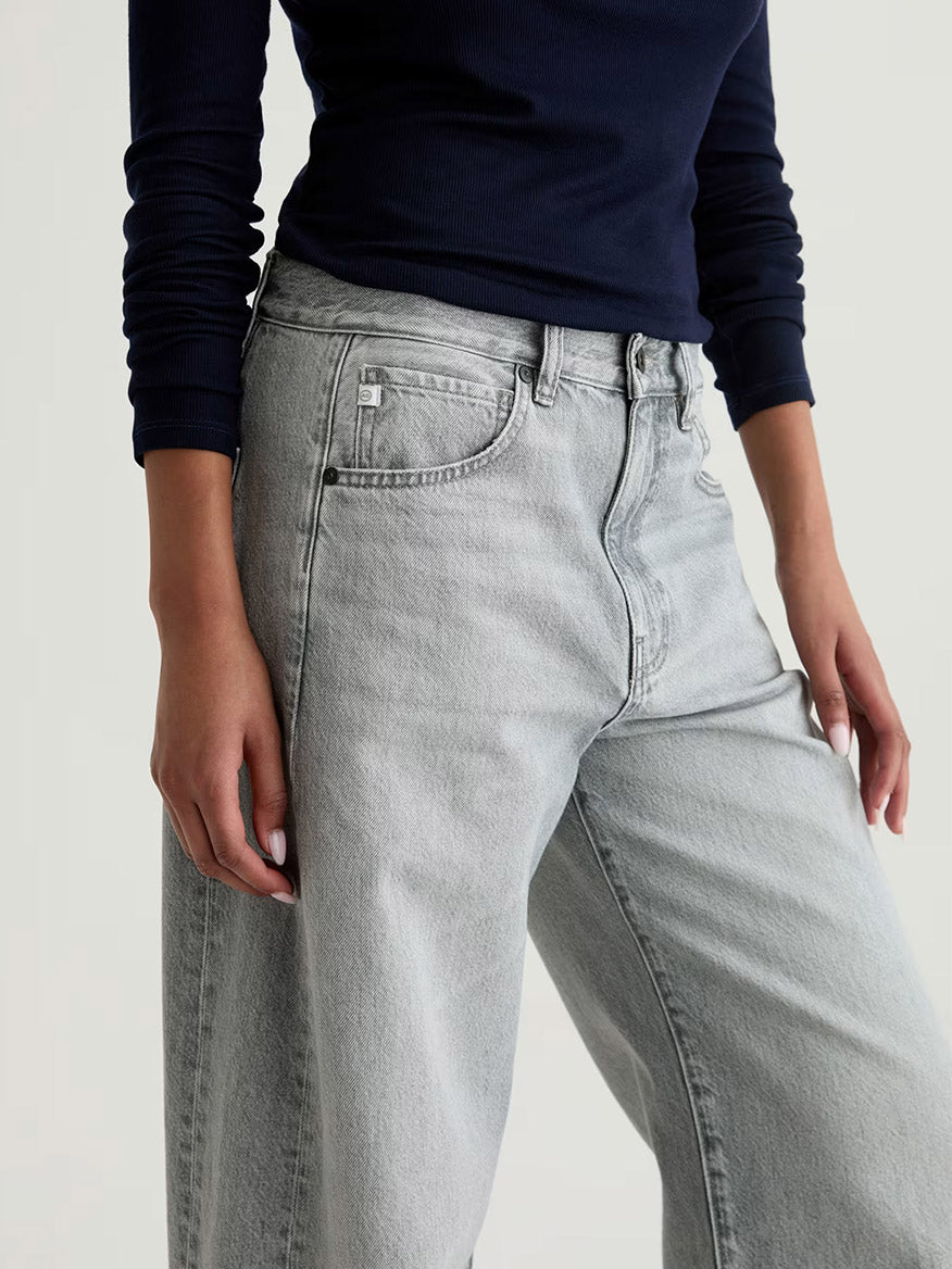 Wearing the AG Jeans Hattie High Rise Barrel in Al Fresco, a person is shown from mid-torso to mid-thigh in a navy long-sleeve top and light-wash non-stretch denim jeans against a plain background.