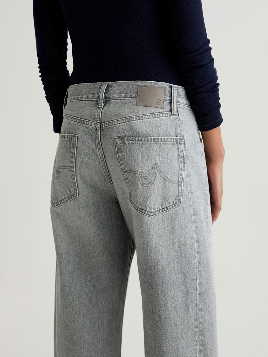 Person wearing AG Jeans Hattie High Rise Barrel in Al Fresco—light gray, non-stretch denim jeans—with a long-sleeve navy top, photographed from the back against a plain background.